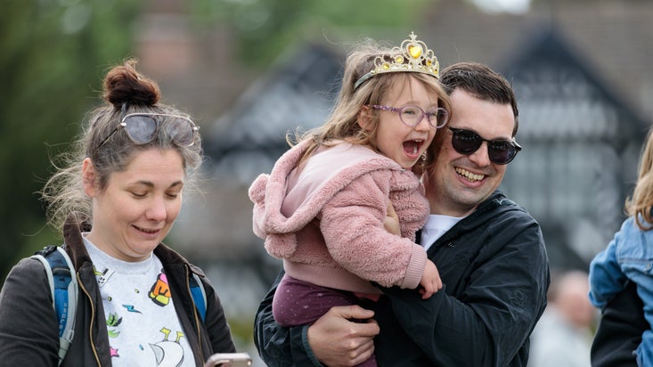 Two adults outdoors, one holding a smartphone and the other carrying a child in a toy crown. Background includes greenery and a blurred building.